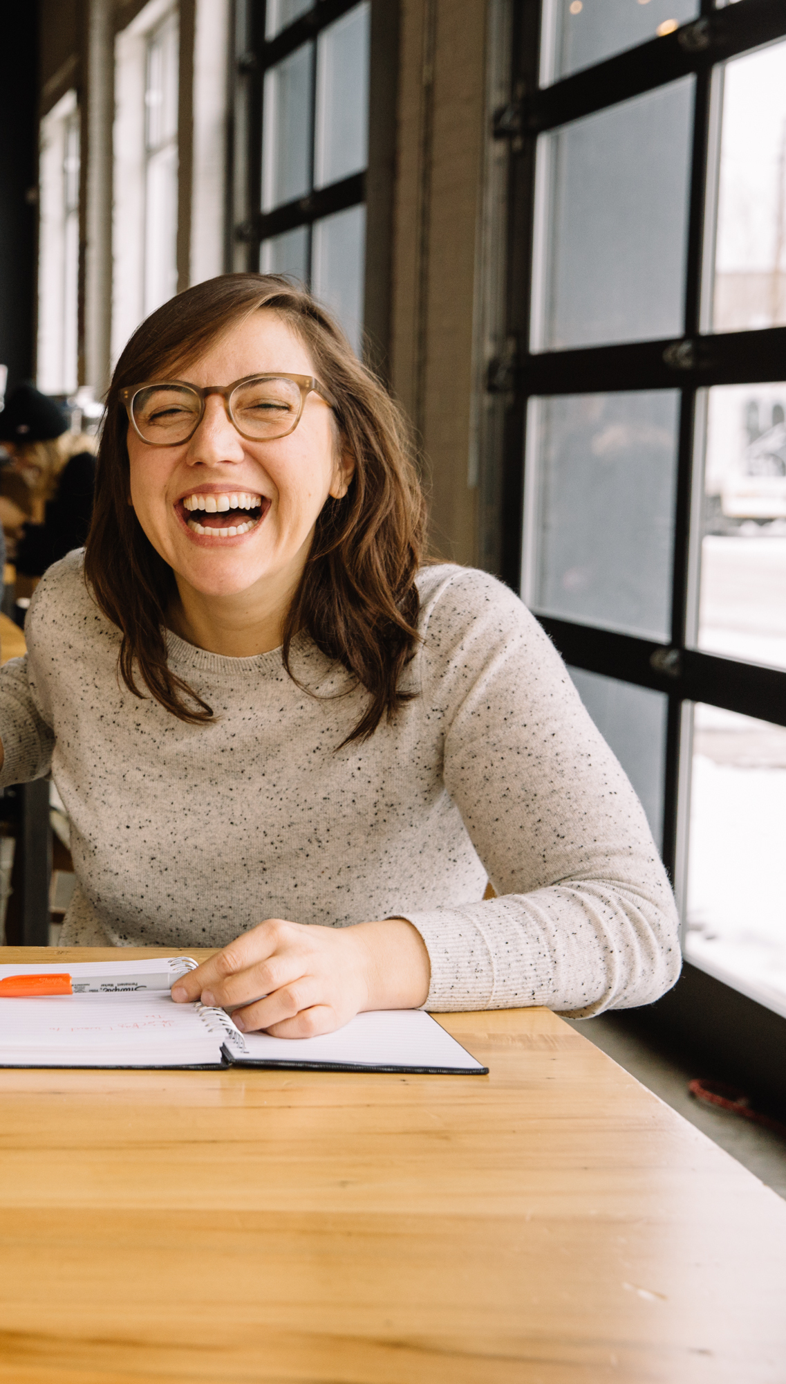 A photo of author Naseem Hrab laughing. She is sitting at a wooden table with a notebook open in front of her. A window is on her left.